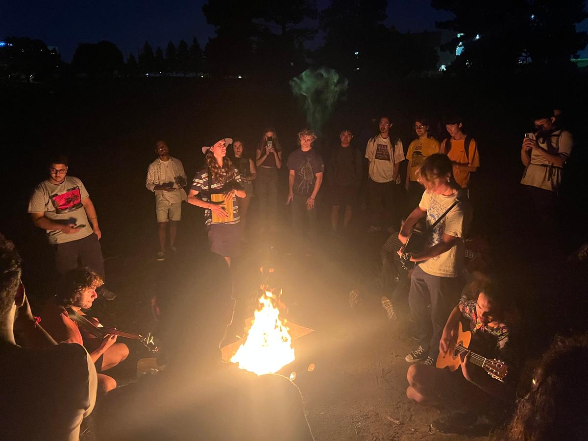 Crowd gathered around campfire with some playing guitar.