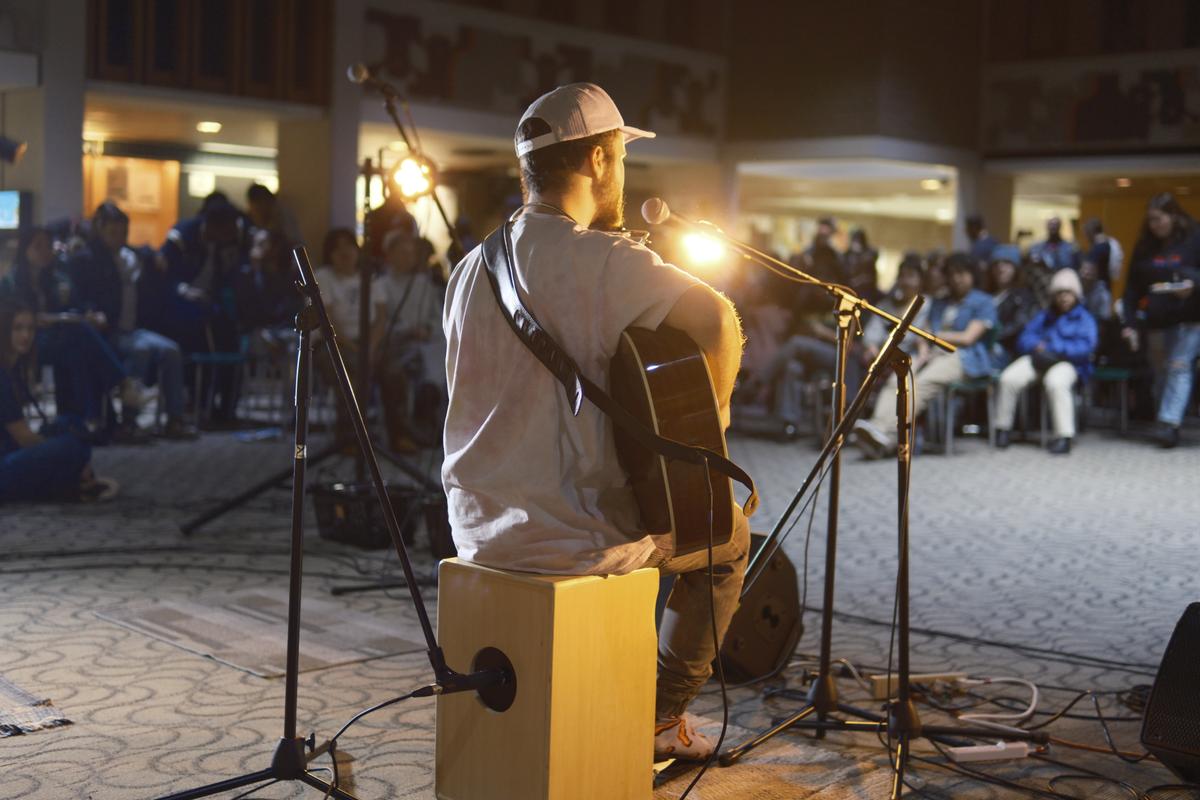 Person sat on cajon playing acoustic guitar and singing to an audience.